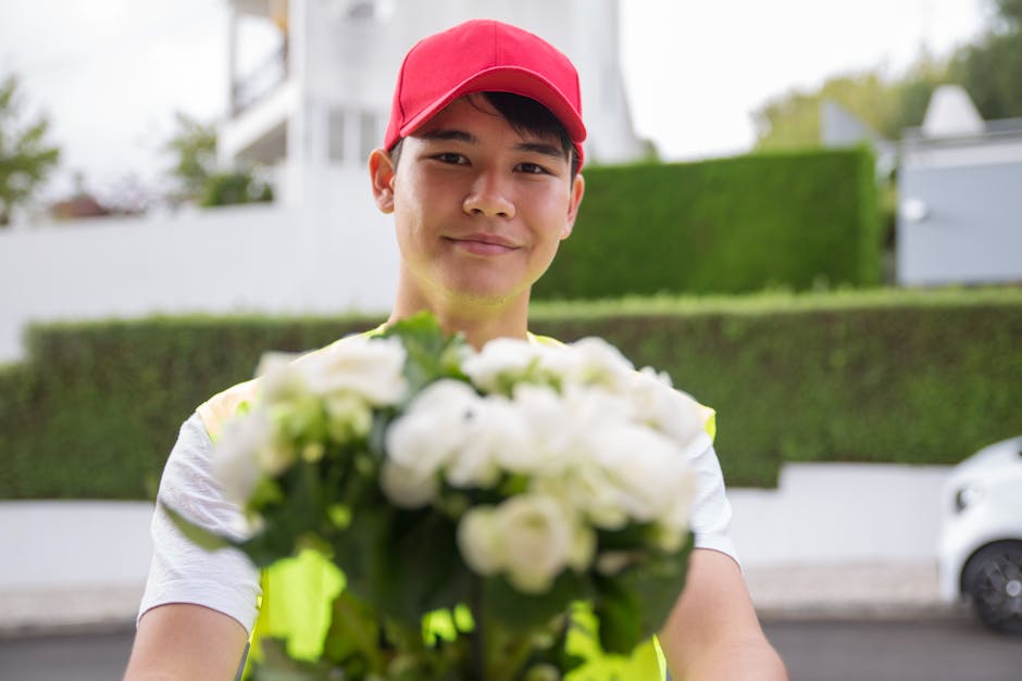 A delivery person wearing a red cap and a bright yellow vest extends their arm forward, holding a bouquet of fresh white flowers with green foliage. The bouquet is wrapped in transparent plastic and i