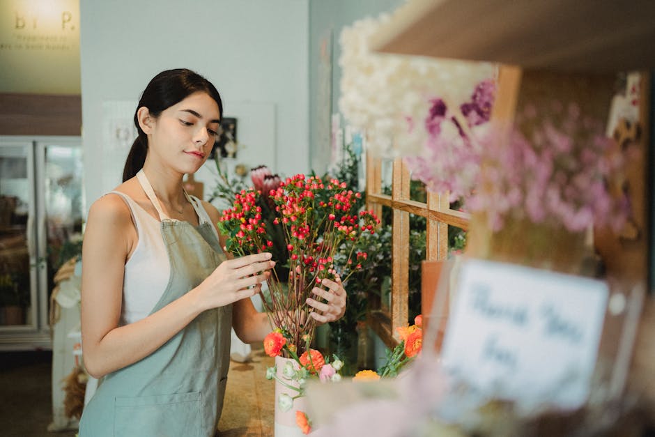 A young woman with dark hair, dressed in a white sleeveless top and beige apron, is selecting a bouquet of small, vibrant pink and orange flowers with green stems and petals. She stands in a florist s