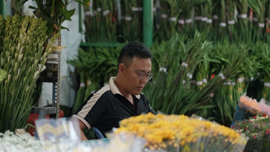 A man with short black hair, glasses, and a dark polo shirt with beige collar and sleeves, is seated at a floral stall in Chinatown. He is focused on arranging or selecting a large bouquet of yellow f