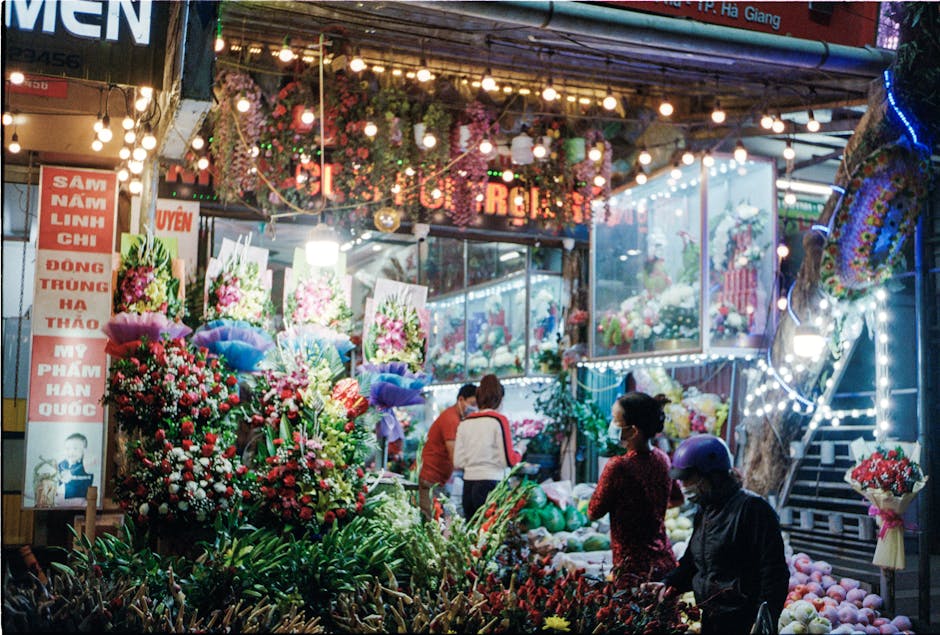 A vibrant flower stall in Chinatown illuminated by multicolored string lights, showcasing an array of fresh floral arrangements in various shades of red, pink, white, and purple. The display includes