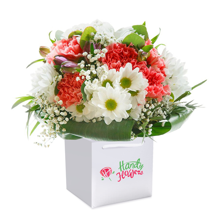 Set against a radiant, white backdrop, this dazzling floral ensemble rises confidently from a modern square gift bag-clean-lined, with sturdy handles and a subtle sense of celebration. The bouquet overflows with sumptuous coral and peach carnations, their ruffled petals soft and inviting, while bursts of red echo the dynamic atmosphere of Chinatown's streets. Crisp white daisies with gleaming chartreuse centres contribute cheerful contrast, recalling the lively windows of nearby bakeries and tea houses. Wisps of delicate white baby's breath weave a gossamer web throughout, creating space and softness between the feature blooms. Hidden within, a few purple-brushed buds suggest both variety and anticipation, reminiscent of the changing seasons around the pagodas on Gerrard Street. Surrounding the flowers, deep green, broad-leaved foliage fans out-supple and glossy, offering a grounding base and amplifying the composition's freshness. The entire arrangement, elegantly contained within the gift bag, signals readiness for swift delivery across London's Chinatown. Whether marking a milestone, brightening a birthday, or conveying appreciation for a neighbor or friend, this bouquet encapsulates the spirit of Chinatown's community: lively, harmonious, and ever welcoming.