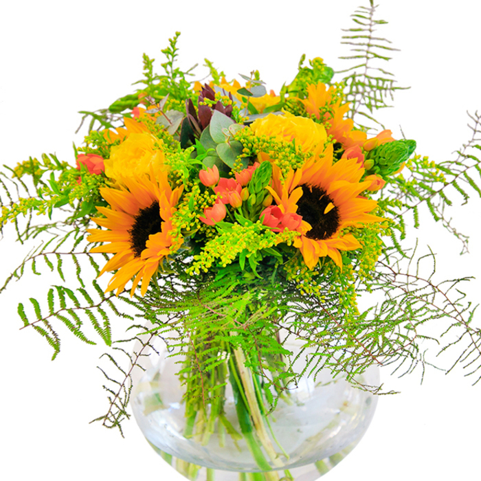 This vibrant bouquet brings to mind a sunlit celebration in the heart of Chinatown. Majestic sunflowers with golden petals and velvety centres fan outwards, their cheerful presence softened by the rounded curves of yellow garden roses nestled nearby, each rose a swirl of subtle fragrance and creamy hue. Interspersed, clusters of red Hypericum berries gleam like festive beads, offering lively punctuation to the gentle sea of yellow and green. Wispy ferns cascade gracefully, their movement akin to the breeze sweeping through Charing Cross Road or drifting through local pagoda gardens. The spherical, clear glass vase presents the submerged stems in their natural, untamed beauty, water shimmering below the blooms in delicate tribute to Chinatown's riverside traditions. Subtle hints of blue-green eucalyptus add a hint of herbal coolness, tying rural textures to the city's vibrancy. Captured in perfect light against a crisp white surface, the bouquet radiates energy and promise-a harmonious centerpiece designed by Chinatown Flowers to mark birthdays, new beginnings, or any moment that calls for a heartfelt dose of joy and celebration within the local community.