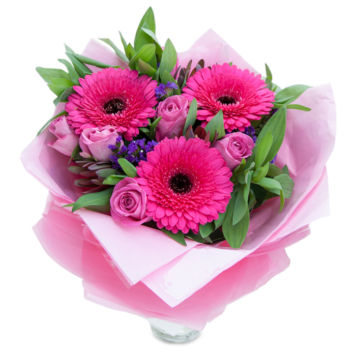 There's a playful grace to this composition that feels right at home among the colourful bustle of Chinatown and the Soho streets nearby. Three vibrant Gerbera daisies in a confident fuchsia hue take centre stage, their layered petals forming a sunburst around dark, contrasting centres that add depth and personality. Surrounding these focal flowers are several soft lavender-pink roses - the kind whose velvety petals sit close in a budlike curl, promising a gentle unfurling over the next days. Sprigs of purple statice peek through the arrangement, offering a delicate, papery texture and a cool, clarifying color note that lifts the warmer pinks. Lush green foliage - broad salal-style leaves with a few slender eucalyptus stems - creates a verdant foundation, the leaves catching light and suggesting a crisp, slightly green fragrance. The bouquet is carefully wrapped: an inner cushion of pale pink matte tissue gives a soft base, while an outer layer of translucent fuchsia-pink cellophane, slightly crinkled, adds a hint of sheen and a cohesive colour statement. The wrapper is folded with an experienced hand, and the clear glass vase rim visible at the bottom signals that the flowers are arranged for immediate display on a bedside table, a shop counter on Shaftesbury Avenue, or a kitchen island in a nearby Covent Garden flat. This piece feels intimate and buoyant - a perfect pick-me-up for birthdays, anniversaries, or a spontaneous 'thinking of you' delivery - and reflects careful floral craftsmanship rooted in local neighbourhood knowledge.