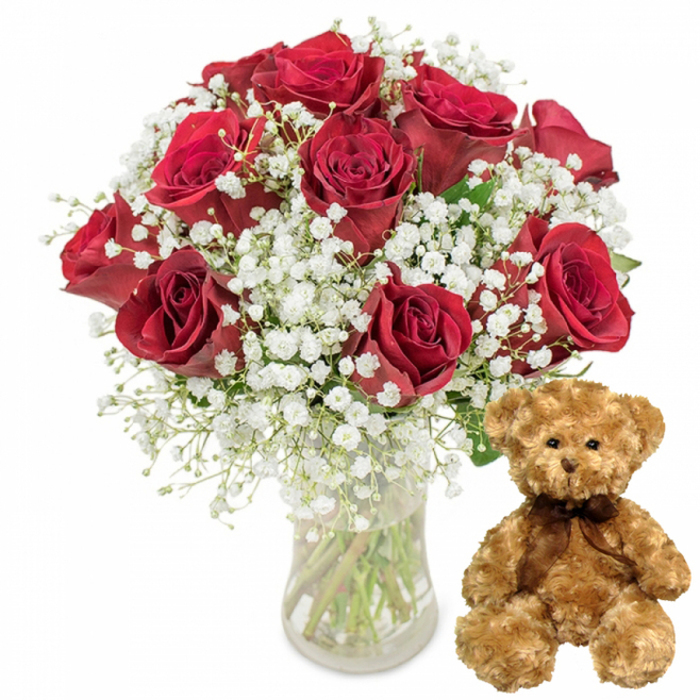 In this bright, minimalist studio scene, a romantic bouquet and a cosy teddy bear are arranged side by side, evoking the feeling of an intimate celebration just off the busy streets of London's Chinatown. On the left, a clear glass vase holds a carefully arranged bouquet of around one dozen deep red roses. The blooms form a rounded, almost sculpted shape, each flower nestled close yet distinct, with velvety petals that curl outward in precise, layered spirals. The colour is a saturated crimson, rich and passionate, catching the light so that subtle highlights and shadows play across the folded petals. Intermixed among the roses, airy sprigs of white baby's breath spread in all directions, their countless minute blossoms giving the arrangement a gentle, floating quality, as though the roses are cradled in a fine white mist. The stems inside the transparent vase are visible in neat, diagonal lines, submerged in clear water that hints at the bouquet's freshness and florist-crafted care. To the right, a light brown teddy bear sits comfortably upright on the same pristine white surface. It has soft, plush fur that appears slightly fluffy, with rounded paws resting forward and small ears perched atop its head. The bear's face is expressive in a quiet way: dark, glossy button eyes gaze ahead, a tiny embroidered nose and subtle stitched smile adding to its sense of warmth and reassurance. Around its neck, a slim dark brown ribbon is tied into a neat bow, giving a polished finish to this comforting companion. With no other objects or background elements to distract, the photograph focuses on this tender pairing-a perfect visual for a romantic flower and teddy delivery from Chinatown to nearby Soho, Shaftesbury Avenue, or Piccadilly, ideal for anniversaries, Valentine's Day, or simply sending love to someone special.