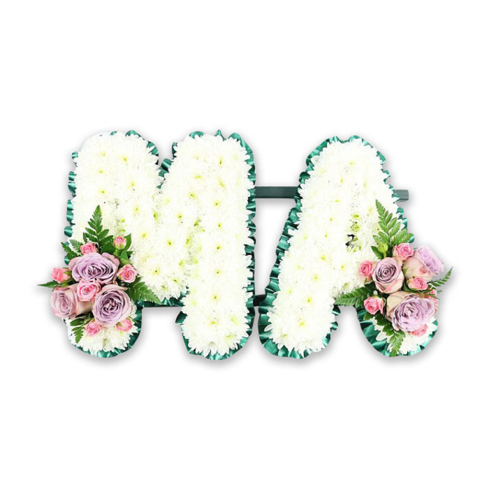 In this carefully lit studio photograph, a bespoke floral tribute spells out the word MA in big, confident capital letters that seem to gently glow against the plain white background. The letters are built on a sturdy base and completely hidden beneath a thick layer of white chrysanthemums. Each flower is fresh and full, with neatly arranged petals forming rounded, cushion-like blooms. Pressed closely together, the chrysanthemums create a dense, almost velvety surface, suggesting both softness and strength. Tracing the outer contours of the M and the A is an emerald green satin ribbon, folded into neat pleats that ripple lightly around every angle. This ribbon adds structure and definition, making the letters stand out clearly from a distance while introducing a subtle sense of movement and craftsmanship up close. The two letters are joined by a slim dark green bar at their base, ensuring they sit as one cohesive design on the display stand. At the lower left base of the M, and balanced at the lower right base of the A, sit two pastel-toned flower clusters. These are made up of lilac roses with cool, dusky undertones, delicate pale pink roses, and tiny pink spray roses, their petals unfolding in soft spirals. Slender fern fronds and small sprays of foliage extend from within the clusters, breaking the straight lines of the letters and adding a soft, organic touch. The tribute feels personal and intimate, ideal for remembering a mother or mama within the multicultural setting of London's Chinatown, perhaps resting beside incense and family offerings. It captures a sense of respect, remembrance, and quiet love, thoughtfully designed for a farewell where flowers speak when words fall short.