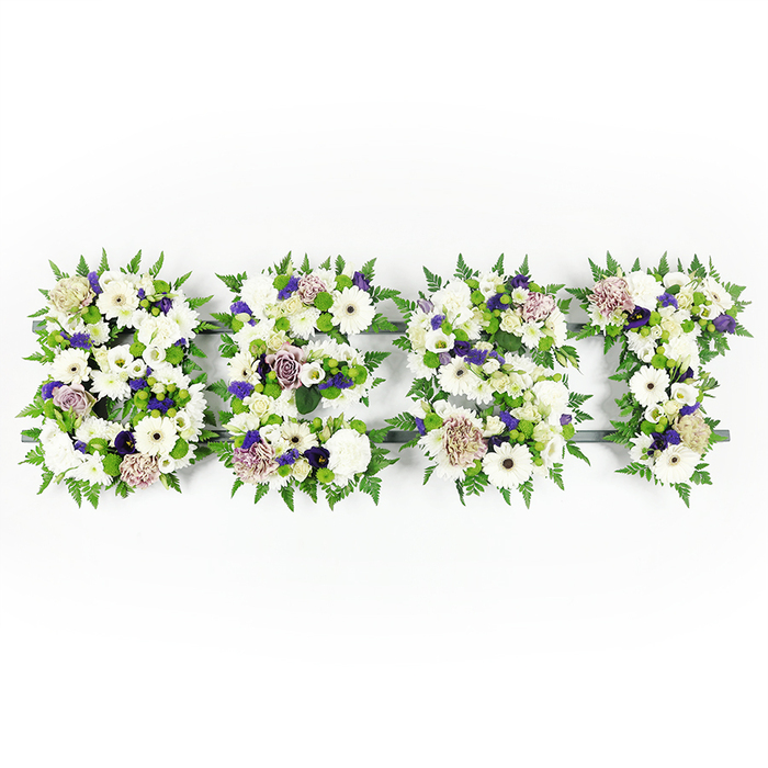 A meticulously formed horizontal tribute spells BEST across a bright white background, each capital letter a compact garden of white and lavender blooms ringed by lush greenery. Large, pure white gerbera daisies dominate the composition with their open faces, accompanied by soft white lisianthus and full, pillowy carnations that convey peace and gentle remembrance. Interspersed among these are soft mauve and lavender roses and deeper violet blossoms which give the letters a measured solemnity and visual depth. The edges of each letter are richly traced with feathery fern fronds, their fine texture creating a halo that both contains and softens the hard edges of the lettering. Small green button chrysanthemums are peppered through the floral masses, providing bright, fresh contrast and a lively, tactile element against the smoother petals. The flowers are anchored to a low, dark structural base that remains barely visible in places, shaping and supporting each character while allowing the blooms themselves to read clearly from a distance. Under even, neutral lighting the whites glow quietly and the purples recede into respectful accent - an aesthetic well suited to funeral chapels or memorial halls. This is the kind of bespoke floral lettering often requested for wakes, services or commemorative displays, crafted with an attention to proportion and lasting freshness. I imagine it set near the Chinatown gate or on a stand at a service on Shaftesbury Avenue, a personal, elegant way to mark a life and offer comfort to those gathered.