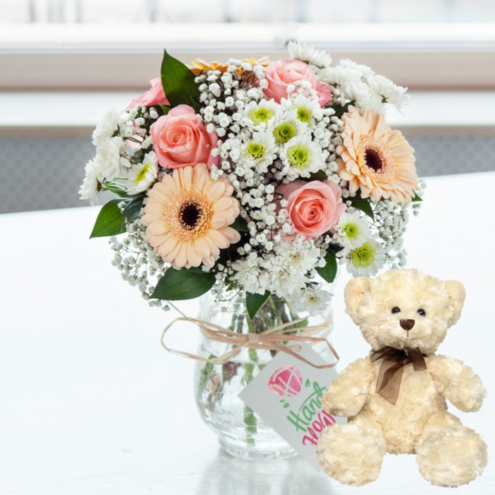An inviting anniversary gift display shows a pastel-toned bouquet placed beside a soft teddy bear on a clean, glossy white surface, as though a florist in Chinatown has just completed a special order. The bouquet, arranged in a simple clear glass jar-style vase, is compact and rounded, with each flower carefully positioned. Blush pink roses, with their petals curling outward in soft spirals, mingle with peach gerbera daisies whose deep brown centres draw the eye. Between them, airy sprigs of white baby's breath and small white chrysanthemums with vibrant lime-green cores add lightness and texture, creating a cloud-like dome of flowers that feels both delicate and joyful. The stems are visible through the glass, anchored in clear water, and a length of natural twine is wrapped around the rim of the vase, tied into a tidy bow. Suspended from the twine is a small, white rectangular gift tag, printed with a stylised pink heart emblem and the word "Handmade" in elegant green script, suggesting a carefully crafted, bespoke arrangement ideal for anniversaries and romantic gifts in central London. To the right, a cream-beige teddy bear sits upright, turned slightly toward the bouquet as if sharing in the sentiment. Its plush fur looks fluffy and comforting, while dark bead eyes, a small brown nose, and a slender brown satin ribbon tied neatly around its neck echo the warm tones of the gerbera centres. Behind them, the background fades into a bright, softly blurred wash of light that hints at daylight streaming through a window overlooking Chinatown's busy streets, yet keeps the focus firmly on the gift. The scene conveys tenderness, reassurance, and celebration, capturing the charm of presenting flowers and a cuddly bear to mark a cherished anniversary in the heart of the city.
