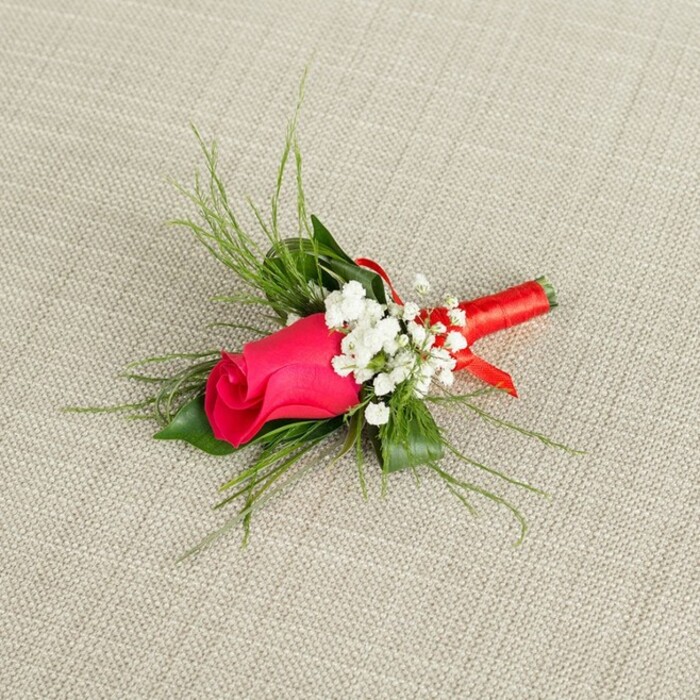 A romantic single-stem boutonnière is displayed at an elegant angle on a softly textured, light beige fabric, its simplicity and refinement evoking a groom preparing for photographs near the colourful arches of Chinatown. At the heart of the design sits a tightly furled rosebud in a dazzling fuchsia, leaning toward deep crimson at its core, the petals smooth and velvety with a subtle satin sheen that suggests they have only just opened enough to hint at the fullness within. Encircling this central bloom are delicate clusters of white baby's breath, each tiny flower head forming a gentle constellation around the rose, softening its intensity and adding a whisper of vintage romance, like lace edging on a wedding dress. Emerging from behind, slender, feathery green sprigs fan outward, reminiscent of asparagus fern or airy ruscus, their bright green tones offering a fresh, lively contrast to the saturated pink and clean white. These wisps of foliage create a sense of movement and natural grace, as if the boutonnière has captured a snippet of a garden hidden behind one of Chinatown's quieter side streets. The stems are carefully aligned and firmly wrapped in a rich, red satin ribbon, the fabric smooth and lustrous, coiling neatly around the base and ending in a small, polished finish that feels both modern and timeless. The ribbon's colour harmonises with the rose while subtly nodding to traditional red accents often seen in local celebrations and tea houses nearby. Even, bright lighting reveals every texture - from the fine fibers of the cloth backdrop to the crisp edges of each petal - conveying a mood of poised anticipation, perfectly suited to weddings, proms, or elegant formal events across central London.