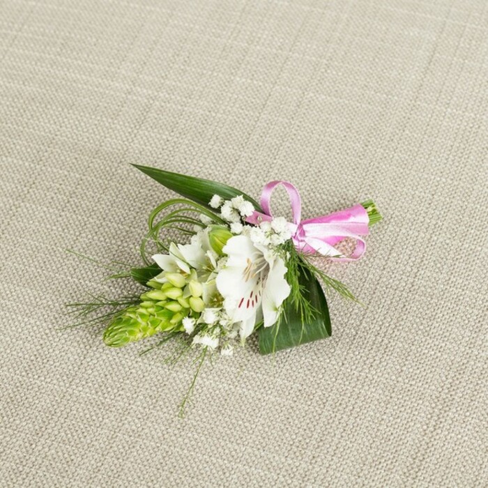 Set against a softly textured, light beige fabric that resembles fine linen, a meticulously crafted boutonniere or small corsage is positioned in the centre of the frame, its compact design radiating quiet elegance. The arrangement's main feature is a single white alstroemeria bloom, also known as a Peruvian lily, its open petals broad and gently ruffled, with slender reddish-brown speckles drawing the eye towards the flower's throat. The stamens are visible, suggesting a blossom at perfect maturity, full of life yet still fresh enough to carry a faint, green-sweet scent. To the left of this focal bloom rises a lime-green spike of tightly clustered buds, likely Ornithogalum or a similar variety, each bud nestled closely to the next to create a conical, textured shape that contrasts beautifully with the smoothness of the lily petals. Interwoven through the flowers is a mist of delicate white baby's breath, their tiny blooms dotting the composition like soft stars and lending a light, romantic haze that suits special occasions across Chinatown and the nearby West End. Underpinning the flowers is a foundation of rich foliage: broader, dark green leaves with pointed, sword-like shapes provide structure, while trailing, grass-fine strands curl and weave around the piece, adding movement and a more natural, garden-picked feel. On the right side, the stems are gathered and neatly wrapped, then finished with a glossy pink satin ribbon tied into a precise yet playful bow, the colour adding a joyful note without overwhelming the calm palette of white and green. The lighting is gentle and evenly distributed, allowing every vein, petal, and ribbon fold to be seen clearly, as though the piece has just been set down by a florist preparing wedding party flowers for delivery from Chinatown Flowers.