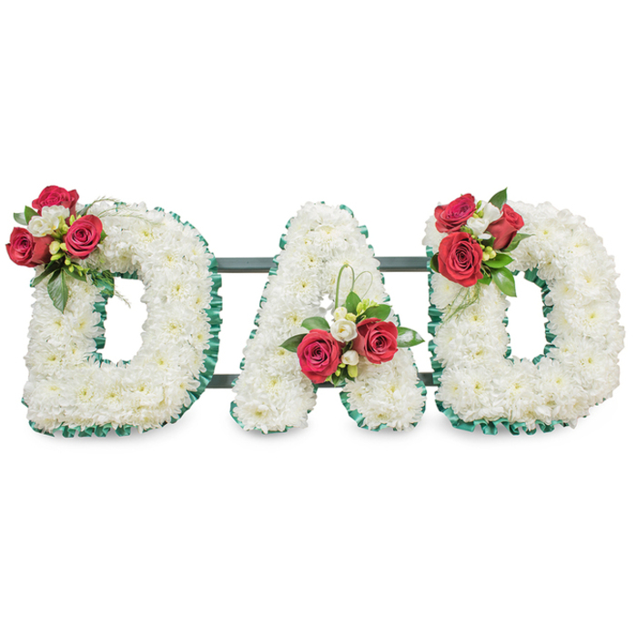 A poignant floral memorial spelling the word DAD in large, block letters, each letter densely covered with hundreds of small white chrysanthemum blooms that together form a uniform, soft-textured carpet of white. The perimeter of each letter is outlined with an emerald green pleated ribbon, its colour and texture creating a crisp frame that sets the white blossoms apart from the bright background. On the left-side D, within the upper curve, a focused spray of open deep red roses sits with petite white freesia and rich dark foliage; the centre A carries a mirrored cluster near the base of its left stem, while the right-hand D echoes the first, creating visual balance and repetition. Subtle strands of glossy green grass thread through the arrangements, offering a modern twist to a traditional sympathy design. The letters are mounted on slender dark green or black metal bars running horizontally just behind the flowers - they're discreet but essential, ensuring the trio remains steady when placed at a service or graveside. Photographed in soft, neutral lighting against a clean white surface, the image emphasises the purity of the chrysanthemums and the contrasting warmth of the red roses, conveying solemnity, love, and remembrance. As a local florist I imagine this tribute being chosen by families in Chinatown and the surrounding West End when seeking a personal, respectful way to honour a father, suitable for a memorial at a nearby ceremony or a quiet graveside farewell.