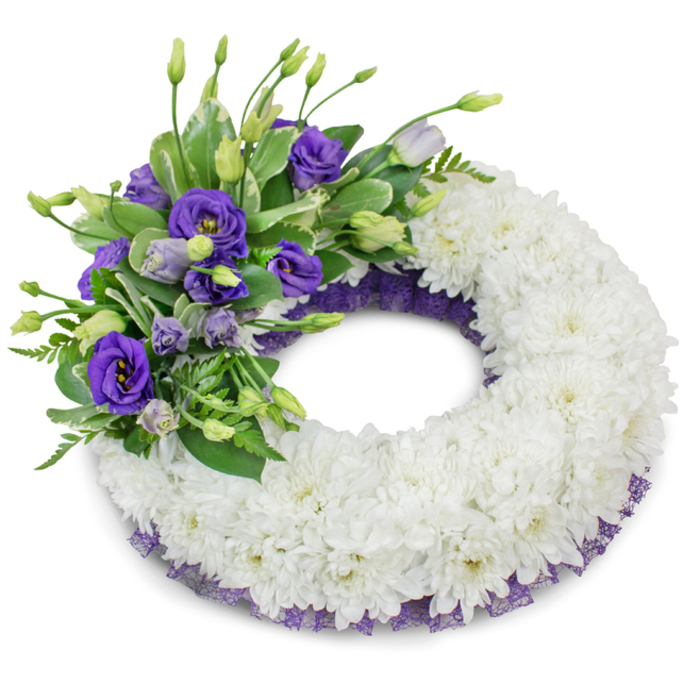 A beautifully balanced circular funeral wreath, photographed against a stark white background so each element reads with clarity and restraint. The bulk of the design is a generous, continuous ring of small white cushion chrysanthemums: densely arranged, their tightly layered petals create a soft, cloud-like texture that conveys serenity and reverence. Positioned on the upper-left quadrant is an elegant accent cluster of purple Eustoma (lisianthus), showing a range of tones from rich violet to pale lavender; open blooms reveal layered petals while unopened buds and upright stems add vertical rhythm and hopefulness. Interspersed among the purple blooms are varied greens - broad, slightly variegated leaves, glossy sprigs, and delicate fern-like foliage - which introduce cool freshness and a natural counterpoint to the florals. A subtle border of dark purple mesh or lace-like ribbon peeks along the inner and outer rims beneath the chrysanthemums, providing a quiet, complementary frame that echoes the lisianthus' hues. The studio lighting produces a soft, diffused shadow under the wreath, giving it gentle prominence and a sense of three-dimensional form. In the context of Chinatown, with its nearby theatres on Shaftesbury Avenue and the calm pockets near Soho Square, this wreath is designed to read as composed comfort-a refined sympathy tribute that offers texture, subdued colour and a measured scent appropriate for funeral services, memorial displays, or moments of private remembrance.