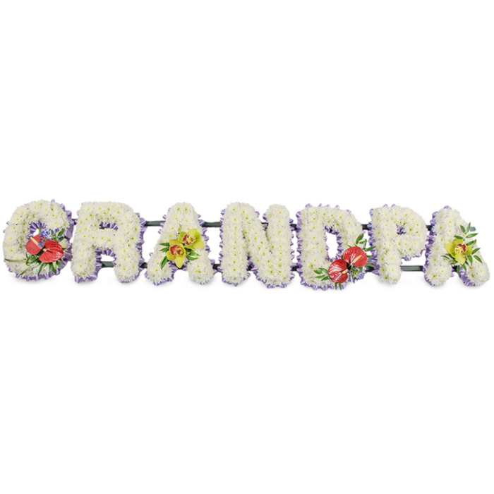 A personalised funeral arrangement spells the word GRANDPA in large, block capital letters, photographed cleanly against a plain white background so the form and detail of the flowers can be clearly appreciated in the mind's eye. Each letter is three-dimensional and robust, its front surface completely covered with small white chrysanthemums laid so closely together that no gaps of foliage or base can be seen. The result is a beautifully even, soft-textured surface, like a series of floral cushions lined up side by side. At the base of each letter, a slim border of pale lavender or light purple ribbon traces a delicate outline, quietly emphasising the shape of the word and adding a gentle touch of colour that complements the white mums. Dark green bars run behind the letters, barely visible yet essential, linking the tribute into a single, continuous piece that could be displayed on a stand during a funeral or memorial in Chinatown. Four of the letters carry carefully designed sprays to introduce colour and meaning. The 'G' and the 'D' are accented with rich red anthuriums, their polished, heart-shaped spathes and central spadices standing out vividly against glossy green foliage and touches of subtle filler flowers, expressing devotion and deep remembrance. The first and final 'A' feature soft sprays of yellow-green orchids, their refined, star-shaped blooms and buds nestled into fresh leaves to suggest grace, admiration, and enduring love. The tribute feels serene yet personal, very much like something a local Chinatown family might choose to honour a grandfather who held an important place at the heart of the home, offering one final message shaped in flowers.