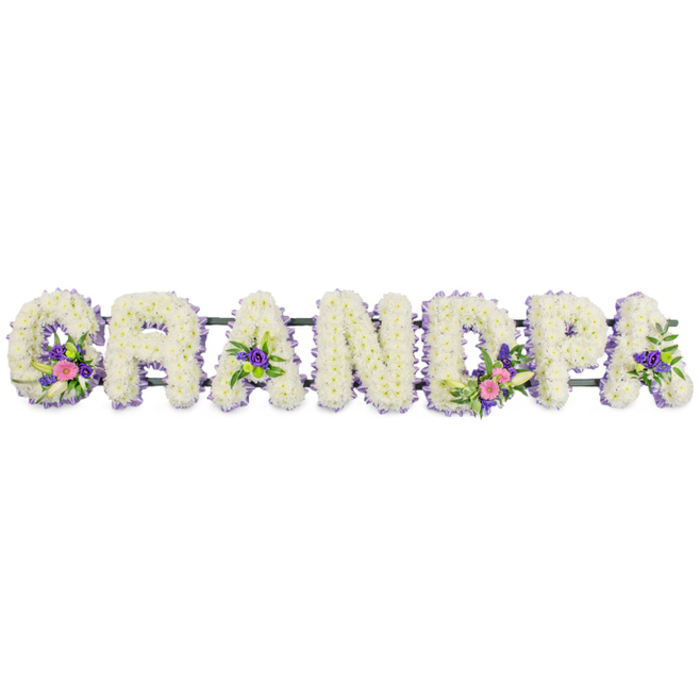 A custom floral name tribute in the photograph spells out GRANDPA in seven bold, flower-covered letters arranged horizontally, perfectly aligned against a crisp white background that gives the tribute a clean, contemplative feel. Each letter is entirely faced in compact clusters of small white chrysanthemums or similar multi-petaled flowers, arranged so tightly that they create a rich, cushion-like surface reminiscent of soft fabric. Along the outer edges, a meticulous line of tiny lilac and purple flowers-possibly statice, heather, or similar filler blooms-frames the white centres, adding a quiet contrast and helping the letters stand out clearly. The florist has added thoughtfully composed accent sprays to certain letters: the G features a small bouquet of vibrant pink and fuchsia blooms, offset by a single elegant white lily-shaped flower and fresh green foliage; the first and last A contain more modest clusters of purple flowers amid dark green leaves; and the D presents another showpiece grouping mirroring the G, with its blend of pink, white, and purple tones. The letters are mounted on a slim dark green frame or rail that runs along their back, mostly hidden beneath the flowers but essential for keeping the tribute straight and stable for display in a funeral setting. The overall look is serene, balanced, and filled with affection, the sort of personalised Grandpa arrangement a family in central London's Chinatown might request to honour a cherished patriarch who spent his days around Gerrard Street, Leicester Square, or the nearby lanes. The tribute feels both traditional and deeply personal, a floral expression of memory, gratitude, and gentle farewell.