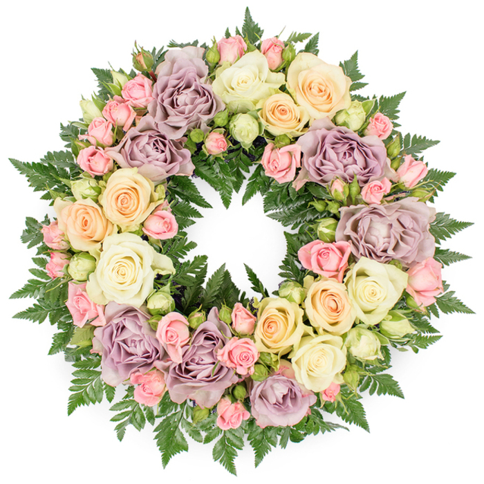 This image shows a beautifully composed memorial wreath placed against a simple white background, allowing every detail of the flowers to stand out. The wreath is perfectly round and richly filled, creating a soft, continuous ring of blossoms that feels both abundant and gentle. Larger roses in creamy white and soft ivory are the central focus, many fully open so that their petals unfurl in generous, rounded layers. They are interlaced with smaller blush pink spray roses, which appear in varying stages of bloom: some tightly closed, others just starting to open, their tiny petals cupped closely around the centre. Threads of soft peach and light buttery yellow roses move through the arrangement, casting a warm, almost candlelit glow among the cooler tones. Distinctive dusty lavender and muted mauve roses punctuate the circle, bringing a refined, slightly nostalgic note that deepens the colour harmony. The flowers sit atop a bed of dark green fern foliage; the ferns' finely serrated fronds peek out around the outer and inner edges of the wreath, their rich colour providing contrast and an organic, garden-like feel. The lighting is even and calm, as though the wreath has been placed in a quiet room before being taken to a funeral service or remembrance ceremony in Chinatown. Without any added decorations, the design speaks purely through the language of flowers, offering a sense of peace, respect, and heartfelt condolence. It resembles the kind of carefully prepared tribute that Chinatown Flowers might hand-deliver to local chapels, temples, or family homes when words alone are not enough to express sympathy.