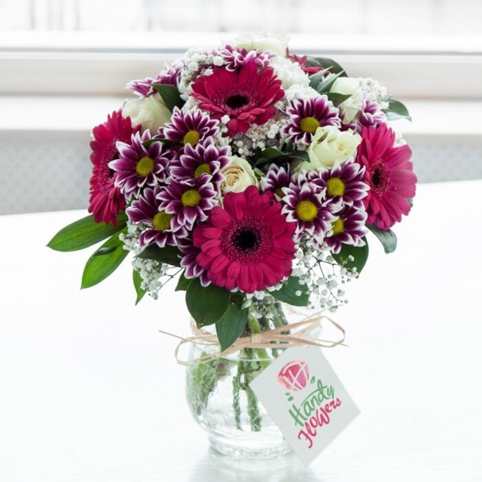 A richly coloured mixed flower arrangement is showcased in the centre of the frame, placed in a low, clear glass vase with a subtle textured pattern that catches the light. Behind it, a bright, softly blurred white background suggests a sunlit window typical of a compact Chinatown apartment or studio, bathing the bouquet in natural glow. The design revolves around several striking gerbera daisies in shades of deep fuchsia and vibrant magenta, their dark, almost ink-coloured centres forming strong, graphic circles that command attention. Surrounding these focal flowers are clusters of chrysanthemum blooms, each petal brushed with intense purple at the tip and fading into clean white toward the middle, framing golden yellow centres that sparkle like tiny lanterns. Interwoven among the bolder flowers are sprays of creamy white roses, some fully open with layered petals curling gently outward, others still in round, promising buds, lending the composition a sense of quiet romance and refined elegance. Airy stems of white gypsophila (baby's breath) drift lightly throughout, softening transitions between the larger blooms and adding an almost cloud-like texture. Lush, glossy green foliage supports the flowers from beneath, with leaves peeking out at varying angles to create structure and depth, as a seasoned florist from Chinatown Flowers might do when preparing an arrangement for delivery across the West End. Through the glass, the straight, freshly cut stems are visible in clear water, reinforcing the impression of just-arrived freshness from a morning run to the flower market. The neck of the vase is encircled with a simple twine bow that introduces a rustic, handmade touch, and from it hangs a small white gift tag printed with a pink rose symbol and elegant script, hinting at a personal dedication for a birthday, Mother's Day, or a heartfelt thank-you on a side street off Wardour Street. The overall mood is joyful yet polished, perfect for brightening a dining table in a Chinatown restaurant or the windowsill of a city flat.