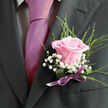 This image offers an intimate close-up of a man's chest and shoulders, dressed in refined formal wear that feels perfectly at home near the theatres and restaurants around Chinatown. His suit jacket is a deep charcoal grey, almost black, with the clean line of the left lapel forming a dark frame for the floral accent it holds. Beneath the jacket, a light dress shirt with a faint checked pattern peeks through, its cool, tidy grid adding subtle texture. A bold, smooth tie in vibrant fuchsia or magenta pink flows down the centre, its satin finish catching soft indoor light, like a ribbon of colour cutting through the muted cloth of the suit. Anchored to the lapel is a handcrafted boutonniere that becomes the visual focal point. A single light pink rose, petals gently unfurling from a deeper blush core to pale, romantic tips, sits proudly at the centre. Around it, clusters of snowy white baby's breath create a misty, lace-like halo, softening the strong lines of the jacket. Slim, bright green blades of foliage rise behind the blooms, adding a touch of movement, as if echoing the leafy calm of nearby Soho Square on a quiet morning. The stems are bound at the base in a deep green wrap, finished with a neat purple satin bow tied just above the breast pocket. The purple ribbon subtly echoes the pink tie, weaving the colours together into a cohesive, modern palette. The photograph captures a moment of poised celebration, perfect for a Chinatown Flowers groom's buttonhole on a wedding day or a special evening event where elegance and heartfelt detail truly matter.