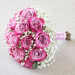 A hand-tied bouquet of roses rests on a pale, linen-like fabric background, the soft texture beneath it reminiscent of a freshly laid tablecloth in one of Chinatown's intimate family restaurants before a celebration. The bouquet is arranged in a gently rounded shape, with approximately twelve roses forming a rich, central cluster of pink tones. Each rose reveals a beautiful gradient: the inner petals are a muted lavender-pink or dusty mauve, lending a cool, romantic undertone, while the outer edges transform into bright fuchsia and deep magenta, delivering a sense of energy and passion. Some blooms remain in tight, sculptural buds, their spirals neat and compact, while others have opened to show layers of velvety petals that curve and twist, adding depth and texture. Framing these roses is a generous scattering of white baby's breath, or gypsophila, whose minute blossoms and slender stems fill the spaces between the larger flowers, creating a delicate, cloud-like halo that softens the entire design. Tiny dark green leaves are just visible underneath, providing contrast and a reminder of the bouquet's natural origins. The stems are carefully gathered and bound at the lower right with a light purple satin ribbon that gleams subtly, giving the handle a polished, comfortable finish for holding during a wedding or engagement ceremony. Over this ribbon, a string of small, pristine white pearls is wrapped in a graceful spiral, recalling traditional pearls often worn at formal occasions. The lighting is soft and even, accentuating the petals' curves and the airy texture of the baby's breath without creating heavy shadows. This elegant arrangement suits modern and traditional weddings alike, making it an ideal image for floral inspiration or for envisioning romantic deliveries throughout Chinatown's vibrant streets and nearby city venues.