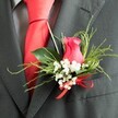 This image softly focuses on a single, stylish boutonniere attached to the lapel of a dark charcoal grey suit, capturing the polished calm before a wedding or special event in London's Chinatown. At its core is a velvety rose bud in vivid cerise-pink, partway between deep pink and fuchsia, its petals curled inward in a delicate spiral that suggests new beginnings. Framing the rose, airy bunches of white gypsophila create a gentle halo, the tiny blossoms dotted like fine lace against the darker fabric of the suit. The greenery is artfully arranged: broad, glossy green leaves anchor the design close to the lapel, while slender, feathery fronds reach slightly outward, giving the buttonhole a sense of movement and freshness, reminiscent of flowers freshly chosen from a nearby Chinatown market stall. The stems are gathered and finished with a bright red satin ribbon, tied into a tidy bow whose rich colour mirrors the celebratory reds that adorn the surrounding streets and lanterns. Just beneath the lapel, a smooth, unpatterned red silk necktie lies flat against a subtly patterned grey dress shirt, the tie's sheen catching the light and tying the whole colour story together. The suit appears to be made from a fine wool blend, its charcoal tone deep and sophisticated, providing a perfect backdrop for the vibrant floral accent. This composition captures the harmony between tailored menswear and floral artistry, ideal for a groom or groomsman preparing to walk into a Chinatown banquet hall, buttonhole perfectly in place, carrying with it a quiet message of love, tradition, and joyful celebration.
