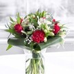 A striking red and white bouquet stands proudly in a clear cylindrical glass vase, its neatly trimmed green stems visible through the fresh water like a cluster of polished bamboo. Deep red roses, their velvety petals just beginning to unfurl, form the romantic heart of the design, joined by a richly ruffled crimson carnation at the front whose layered, frilled petals add a sense of movement and texture. Around them, pure white and creamy blooms - including elegant white alstroemeria with fine speckles at their throats, and several tightly closed white lily buds - bring a feeling of calm balance and promise, as if the arrangement will keep evolving over the next few days on a windowsill overlooking Chinatown's busy streets. Tiny clouds of white gypsophila, or baby's breath, float delicately between the larger flowers, softening the stronger colours and giving the bouquet a light, airy halo. At the base, broad, glossy dark green tropical leaves are carefully curled and wrapped to form a structured collar that cradles the flowers, while slimmer, eucalyptus-like sprigs and pointed foliage weave upwards, adding depth, gentle fragrance, and layers of green. The bouquet rests on a pristine white surface against a softly blurred, bright background, reminiscent of daylight streaming through a shop window on Gerrard Street. The even, natural lighting catches the reds, whites, and greens so every petal appears freshly opened, making the arrangement feel like a warm, thoughtful gesture - ideal for an anniversary dinner at home, a birthday surprise delivered to an office in Chinatown, or simply a beautiful way to send affection and celebration across the neighbourhood.