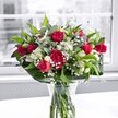 A lush, romantic bouquet of red roses, red carnations, white alstroemeria, and clouds of dainty white gypsophila stands proudly in a clear glass vase on a bright white tabletop, bathed in soft daylight from a large window. Each crimson rose, some fully open with velvety, layered petals and others held in tight, promising buds, becomes a focal point, their deep red tones glowing against the clean, pale background. The carnations echo this richness, their ruffled petals forming rounded, textured blooms that fill out the heart of the arrangement with a sense of abundance and tradition, like something you might see carried along Gerrard Street on a festive evening in Chinatown. Threaded between the bold reds are stems of white alstroemeria, many still in bud, their slender, trumpet-like flowers edged with faint green touches that add freshness and quiet elegance. Tiny star-like blossoms of baby's breath are scattered throughout, creating a light, misty veil that softens the stronger colours and brings a feeling of airiness. Around the base, broad, glossy green leaves form a natural collar, providing structure and a deep, grounding contrast, while slimmer, lighter greenery weaves upward, adding rhythm and movement. Through the transparent vase, the neatly trimmed stems are clearly visible, fanning out in clear water that hints at how freshly this bouquet has been arranged. The background, gently blurred and almost monochrome, allows the bouquet's reds, whites, and greens to stand out with striking clarity, as if the entire scene has paused to honour these blooms. It feels like a bouquet chosen to celebrate love, gratitude, or a meaningful milestone, the kind of gift you might have delivered to a favourite restaurant or apartment in Chinatown to brighten a special day with colour and life.