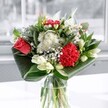 A contemporary glass-vase bouquet stands on a sunlit surface, its colours glowing softly as if catching the light from a Chinatown shop window on a clear morning. At the heart of the arrangement, a single coral-red rose opens in full, its layered petals velvety and plush, framed by a ruffled crimson carnation that deepens the sense of passion. Around these central blooms, clusters of crisp white chrysanthemums and tiny daisy-like flowers float like little clouds, their petals fine and feathery, lending the design a fresh, breath-of-air feeling. Creamy white lilies, mostly in smooth green-tipped buds, rise just above the other flowers, suggesting moments of beauty still to come, like blossoms about to open along Gerrard Street in early spring. Broad, glossy green leaves are carefully folded and arched around the base, creating a polished collar that cradles the bouquet and gives it a modern, sculptural line. The clear glass vase shows the stems gathered neatly together, adding a sense of honesty and simplicity to the composition. Natural light pours across the scene, bright yet gentle, catching on each petal and leaf so that the reds appear richer and the whites almost luminous. The overall mood is one of quiet celebration and thoughtful design, a piece that would feel equally at home on a dining table above a Chinatown restaurant or on a work desk overlooking Leicester Square. You can almost sense a fresh, leafy fragrance with a hint of sweetness, making this arrangement ideal for marking birthdays, heartfelt congratulations, or simply brightening an ordinary day with a touch of London charm.
