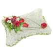 A solemn, hand-crafted sympathy pillow formed from hundreds of small, densely packed white chrysanthemum blooms rests against a clean, white background. The surface reads like a soft fleece of petals: cool, pristine and quietly luminous under gentle studio light, suggesting peace and rest. Around the slightly raised rectangular edge a slim band of light green ribbon peeks through, giving a neat, fresh frame to the cushion and introducing a hint of verdant colour. At the upper left a rich cluster of deep red roses and burgundy carnations gathers like a single, heartfelt gesture; their velvety petals and ruffled textures contrast against the chrysanthemum's fine pom-pom heads. Interspersed among them are small, creamy white freesias (or spray blooms) that catch the eye and lift the composition with delicate points of brightness. Feathery fern fronds and glossy green leaves form a leafy backdrop, while long, arching strands of bright green bear-grass sweep diagonally from the main cluster toward the lower right corner, creating a graceful line and sense of movement across the white field. The arc terminates in a solitary, perfectly formed red rose on the lower right - a poignant focal point that reads as a symbol of enduring love and remembrance. The overall impression is one of dignified tenderness, a classic memorial piece suitable for funerals, wakes or a quiet tribute at a chapel on Shaftesbury Avenue or a service near Leicester Square. Tactile, scented lightly of fresh roses and green foliage, this arrangement communicates comfort, respect and a gentle permanence in moments of farewell.