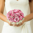 The photograph reveals a beautifully structured bridal bouquet nestled gently in the bride's hands, set against the intricate details of her lace wedding dress. At the centre of the arrangement, blush-pink roses are gathered tightly into a soft, rounded dome, each flower at the perfect stage of openness-full, plush, and delicately spiralled. Their petals display subtle variations of colour, from scarcely tinted blush to a slightly deeper, warm pink, creating a quietly luxurious gradient across the bouquet. Interwoven with these roses, sprigs of white gypsophila, or baby's breath, form a fine, cloud-like border around the blooms, their tiny blossoms scattered like stars across a pale sky. The gypsophila softens the bouquet's outline and adds a gossamer lightness, contrasting beautifully with the denser, velvety textures of the roses. Light drifts over the flowers much as it might fall through the windows of a Chinatown restaurant-turned-wedding-venue, lending a soft glow to the petals and throwing faint, intricate shadows onto the bride's ivory lace bodice. Her gown, rich with lace motifs and delicate embroidery, mirrors the intricate, small-scale detail of the baby's breath, creating a cohesive, romantic visual story. Hints of green foliage can just be glimpsed beneath the blossoms, providing structure and a subtle freshness that enhances the overall palette of blush, white, and cream. The mood is one of serene anticipation, as if the bride is moments away from walking past lantern-strung streets toward her ceremony. You can almost imagine the scent of fresh roses mingling with the aromas of nearby Chinatown bakeries and cafés. This bouquet embodies a refined, classic style, filled with tenderness and intention, designed to be held close through vows, photographs, and the first walk together as a married couple.