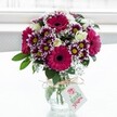 A vibrant mixed bouquet of magenta gerbera daisies, creamy roses, and purple-tipped chrysanthemums stands proudly in a clear, textured glass vase, set against a softly glowing white background that feels like daylight streaming through a Chinatown flat window. The gerberas, in rich fuchsia and deep pink tones with velvety dark centres, create bold, joyful circles of colour, drawing the eye immediately to the heart of the arrangement. Around them, daisy-style chrysanthemums unfurl layers of petals edged in royal purple that gradually fade to white, their golden centres catching the light and adding a lively sparkle. Nestled between these statement blooms are clusters of delicate cream spray roses, some fully open with softly cupped petals, others still in tight buds that hint at flowers yet to unfold, lending the bouquet a sense of gentle romance. Wisps of airy white gypsophila weave throughout the design like a soft mist, giving texture and lightness, while deep green foliage frames and supports the flowers, grounding the brighter hues. The stems are visible through the squat vase, their fresh green lines adding to the sense of just-picked vitality. A simple bow of rustic twine is tied neatly around the neck of the vase, from which hangs a small white gift tag printed with a stylised pink rose and elegant script, suggesting a thoughtful handwritten message for a birthday, anniversary, or thank-you delivered to a home above Gerrard Street or a cosy restaurant off Shaftesbury Avenue. The overall mood is one of cheerful celebration and modern elegance, the kind of bouquet that brightens a grey London afternoon and feels at home in both traditional Chinatown shops and contemporary city apartments.